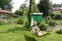 Älterer Mann in einer grünen Hängematte über einem Rasen gespannt, dahinter Gartenhäuschen