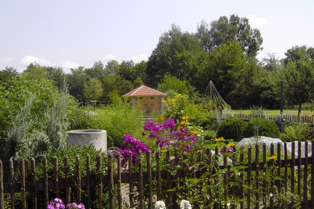 Blick über einen Holzgartenzaun auf Beetbepflanzungen mit Blumen und Gräsern, eine Wasserstelle und ein Gartenhäuschen.