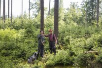 Zwei Förster begutachten einen Wald, einer mit einem Hund, der andere mit einer Axt und Helm