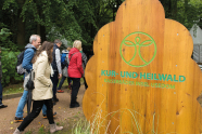 Eine Gruppe Menschen in Regenjacken spaziert an einem Schild aus Holz mit der Aufschrift Kur- und Heilwald vorbei
