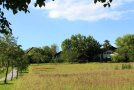 Wiese, im Hintergrund ein Haus zwischen Bäumen, rechts ein weg mit Alleeebäumen