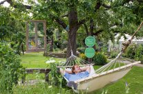 Mann in grüner Hängematte unter einem Baum, dahinter Rasen, Pflanzen, Gartenhäuschen