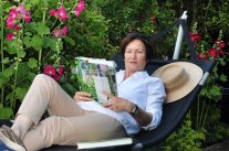 Frau in einer Hämgematte in Gartenzeitschrift lesend vor Stockrosen