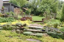 Blick über einen Kräuterrasen durch ein Gartentor auf den Rasen mit Gartenhäuschen