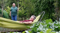 Frau in grüner Hängematte liegend, dahinter steht im Beet ein Mann mit einer Gartenhacke.