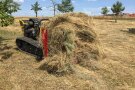 Eine Mähraupe, an welche ein Pick-up-Schwader montiert wurde. Dieser hat bereits eine erhebliche Menge sehr trockenes, langhalmiges Mähgut geladen. Im Hintergrund ist ein Dorf erkennbar.