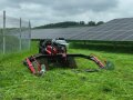 Ein roter Mähgeräteträger mit Raupenlaufwerk mit Doppelmesserbalken. Dieser steht in einem Freiflächenphotovoltaikpark.