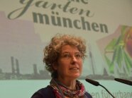 Manuela Barth ist vor der Leinwand, darauf der Schriftzug der Gärten München zu lesen.