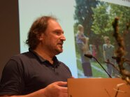 Matthias Schöring steht am Rednerpult, auf der Leinwand im Hintergrund ein Bild mit einer Familie.