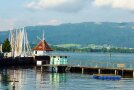 Blick auf einen Steg, mit Häuschen am Ende, viele Segelmasten, im Hintergrund waldige Hügel