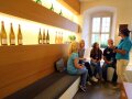 Vier Personen sitzen in modern gestaltetem Raum mit Holz, beleuchteten Weinflaschen; eine weitere Person mit Weinflasche erklärt.