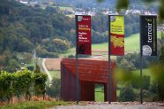 terroir f - Fahnen und sich zur Landschaft öffnendes Tor mit Blick über Weinberg, Felder, Wald und Siedlung