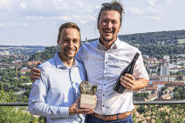 Johannes Burkert und Martin Justus Müller mit dem Award und einer Flasche Wein in der Hand lachen in die Kamera.