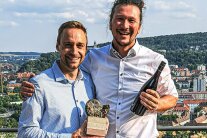 Johannes Burkert und Martin Justus Müller mit dem Award und einer Flasche Wein in der Hand lachen in die Kamera.