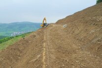 Ein Bagger zieht die schräge Böschung einer Querterrasse ab; der Hang zeigt stufig modelliertes ERdreich
