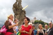 Farbig gekleidete Menschen mit gefüllten Weingläsern in den Händen stehen lachend vor einer Skulptur, im Hintergrund die Festung Marienberg